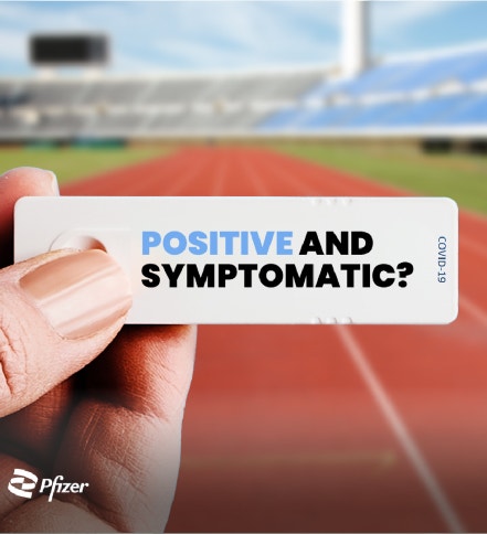 A hand is holding a COVID-19 test strip with the text "POSITIVE AND SYMPTOMATIC?" on it. The background shows a blurred image of a running track in a stadium. In the bottom left corner, there is a Pfizer logo.